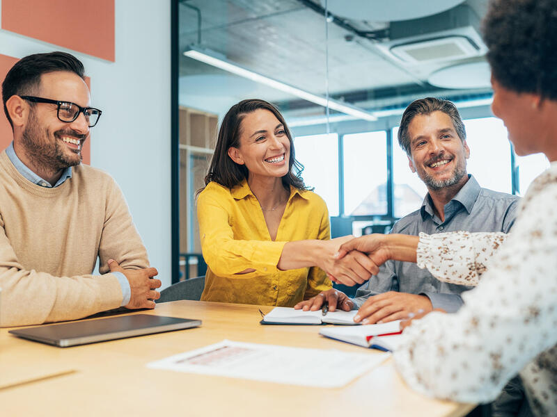 A photo of three people on one side of a table and one person sitting on the opposite side. The person in the middle on the left side and the person on the right side are shaking each others hands. 