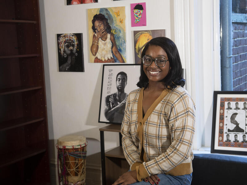 A photo of a woman sitting in a room. Behind her are paitnings and illustrations hung up and a drum sitting on the floor. 