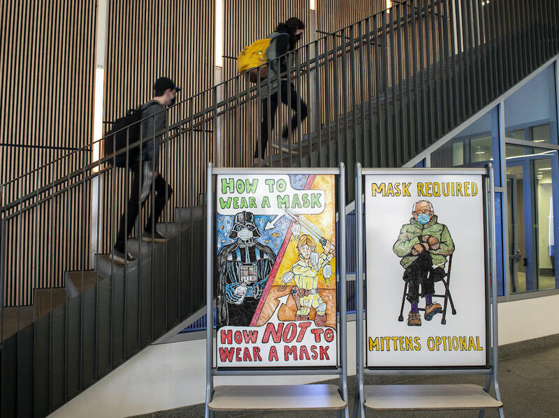 Students walk up the stairs at Cabell Library. A Star Wars-themed drawing on the left whiteboard states \"How to wear a mask. How to NOT wear a mask.\" A drawing on the right whiteboard of Sen. Bernie Sanders states \"Mask required, mittens optional.\"