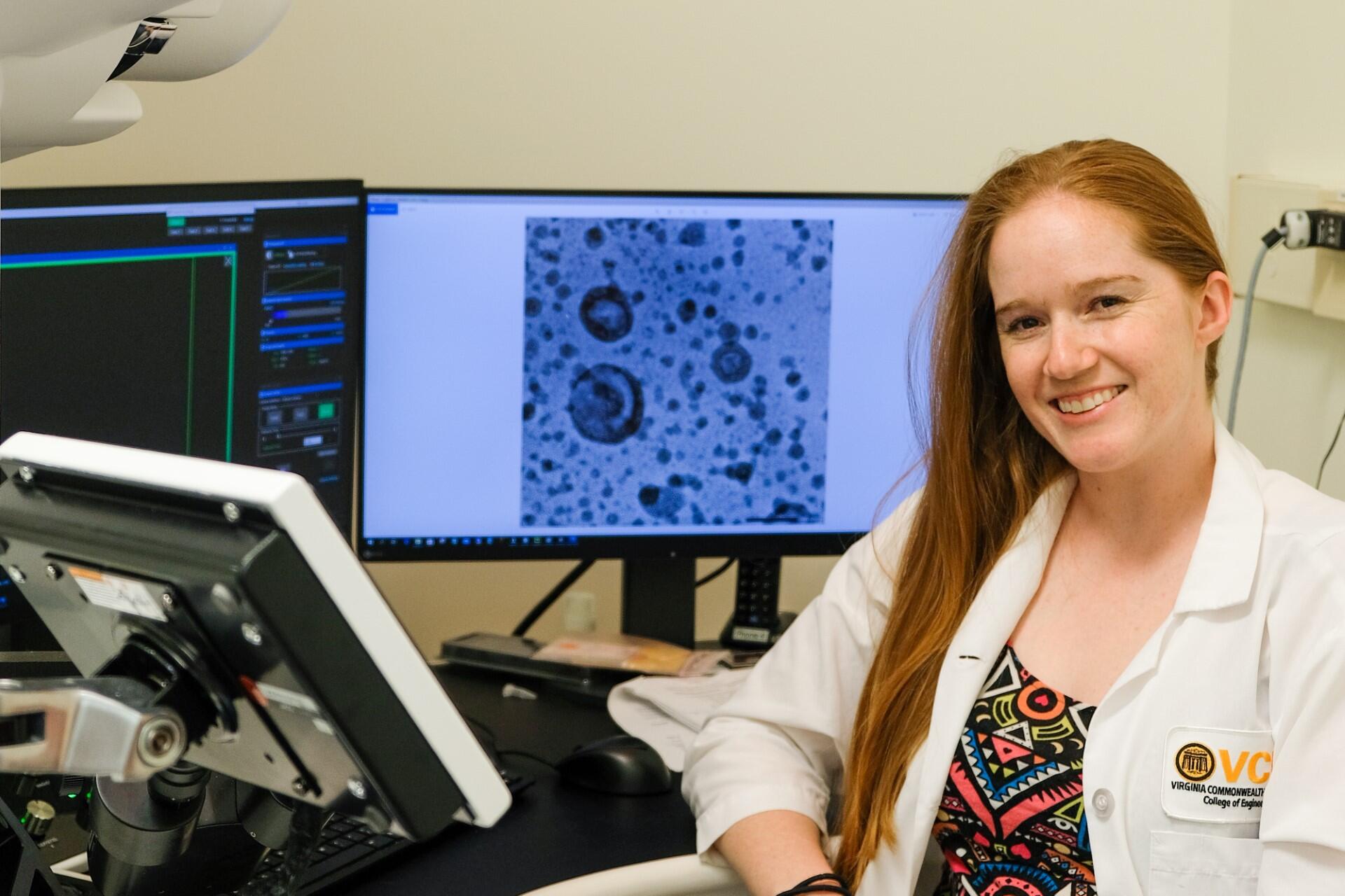 A photo of a woman in a lab coat sitting at a desk with two computer monitors behind her and another screen in front of her. 