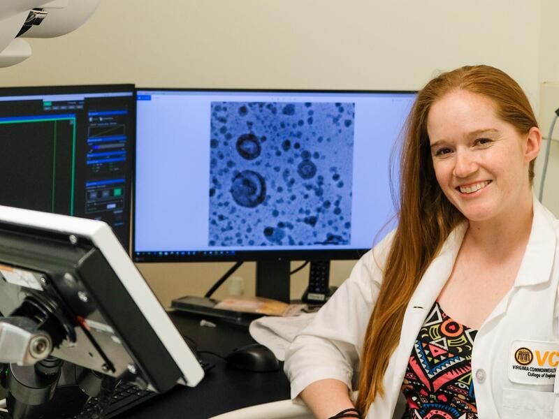 A photo of a woman in a lab coat sitting at a desk with two computer monitors behind her and another screen in front of her. 