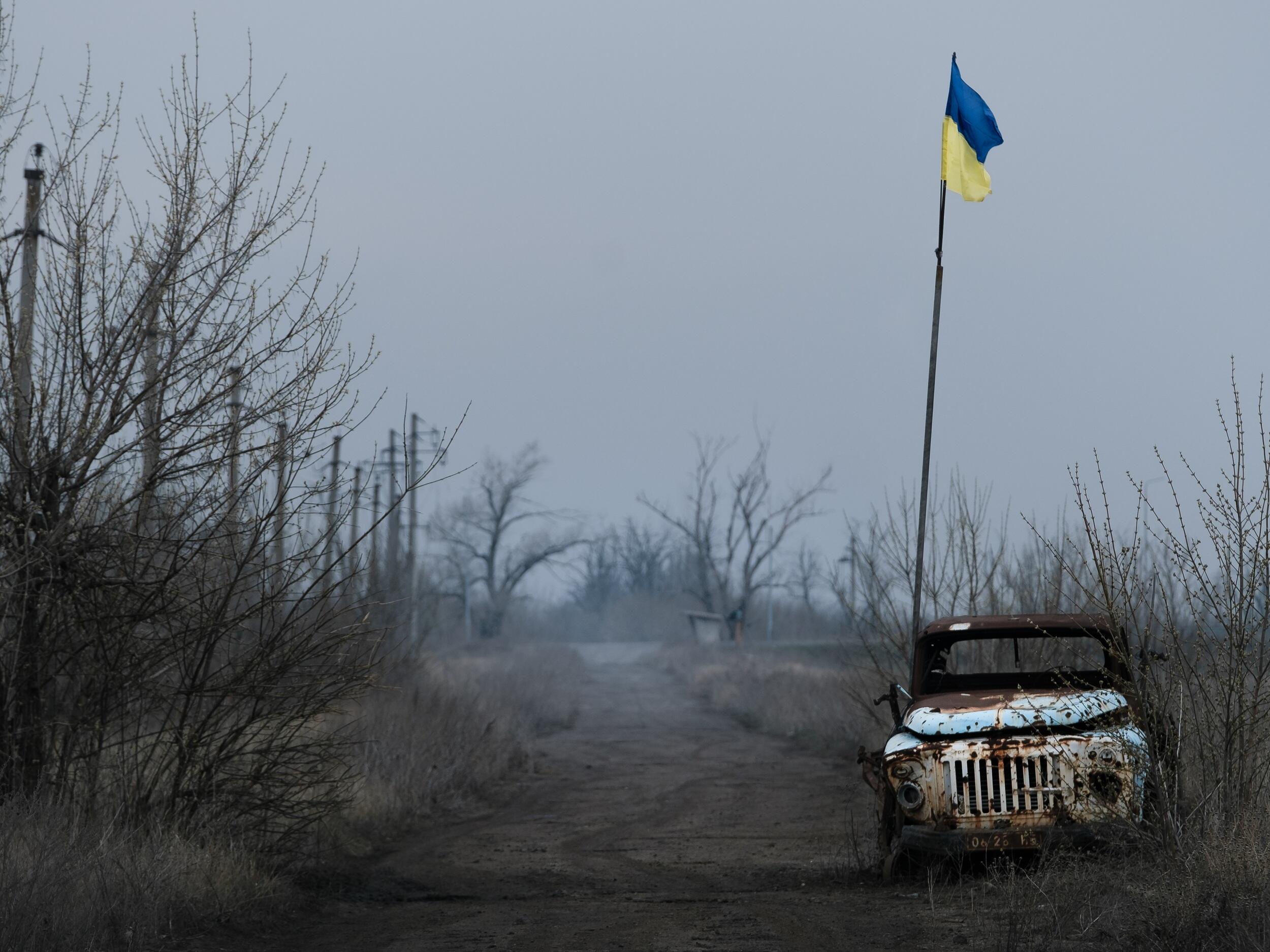A ukrainian flag on a flag pole next to an abandoned jeep in an area with dead trees and a gray sky 