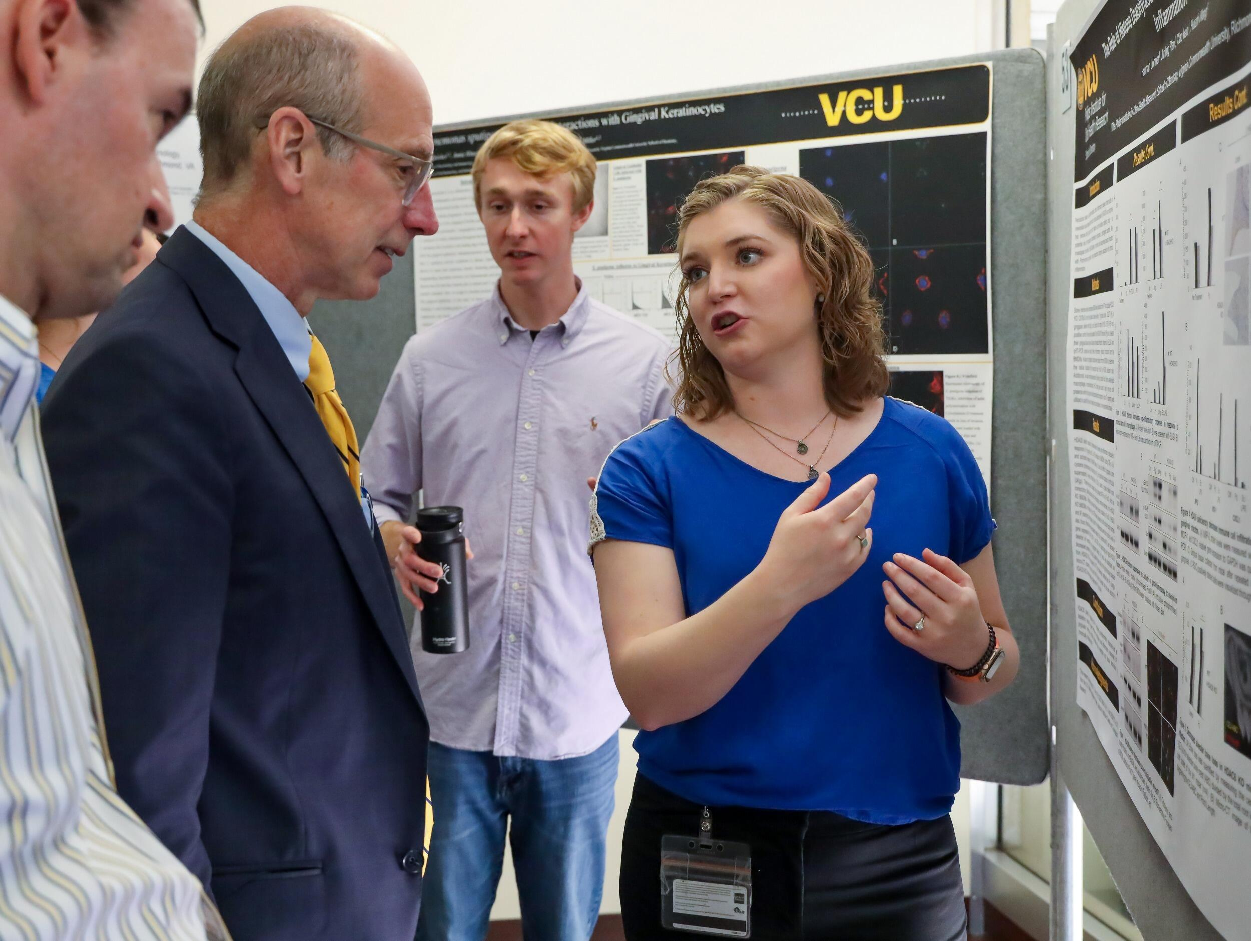 A photo of a woman standing in front of a poster and speaking. To her right three men looking at her and listening. 