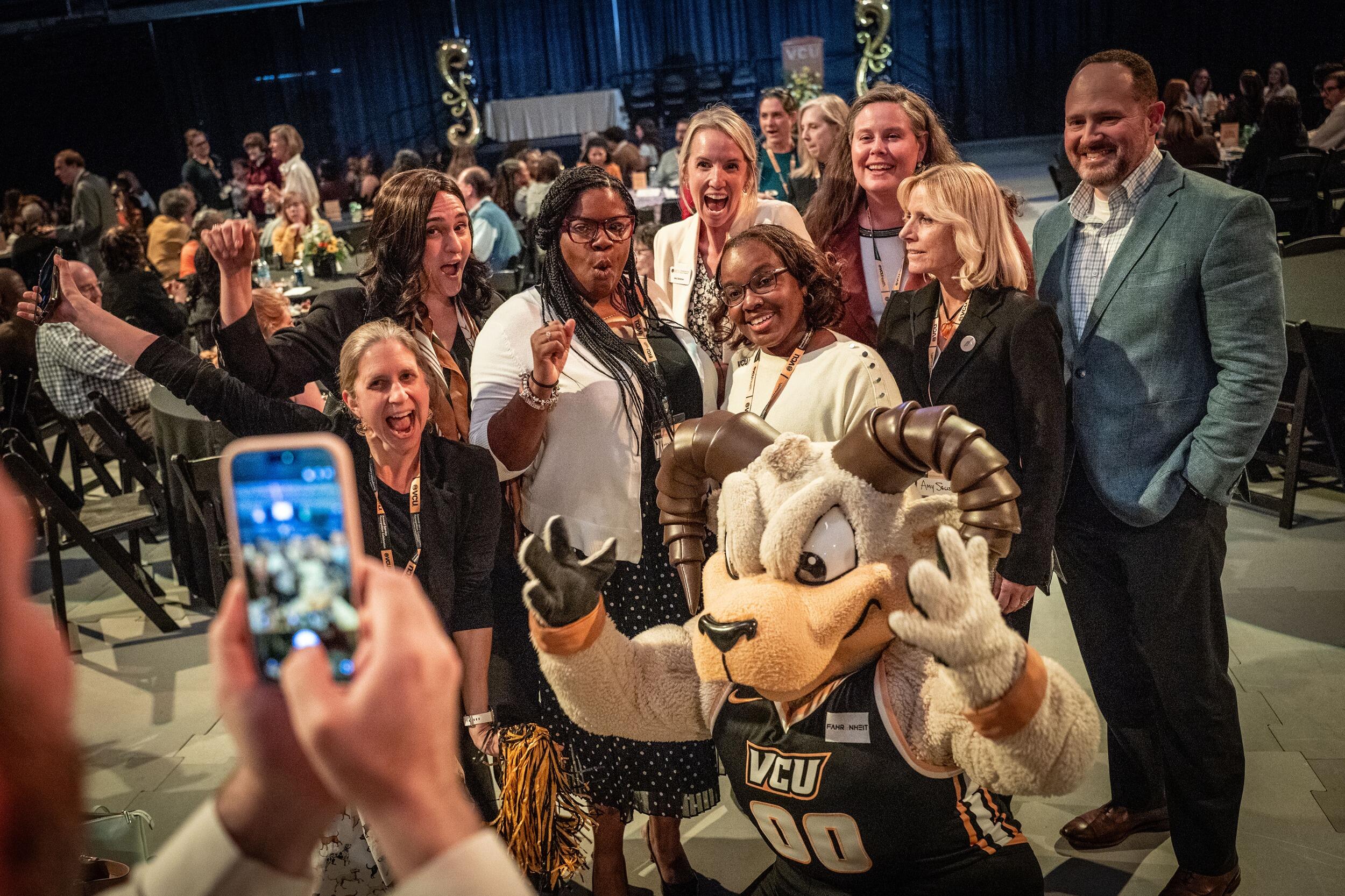 A photo of 8 people posing with Rodney the Ram and one person taking a photo of them with a smart phone. 