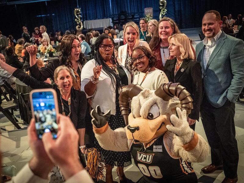 A photo of 8 people posing with Rodney the Ram and one person taking a photo of them with a smart phone. 