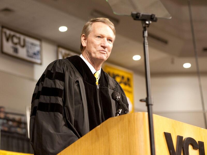 A man in a graduation gown speaks at a lectern.