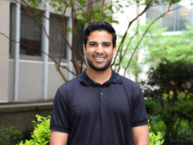 A photo of a man from the chest up standing outside in front of a small tree and some bushes. 