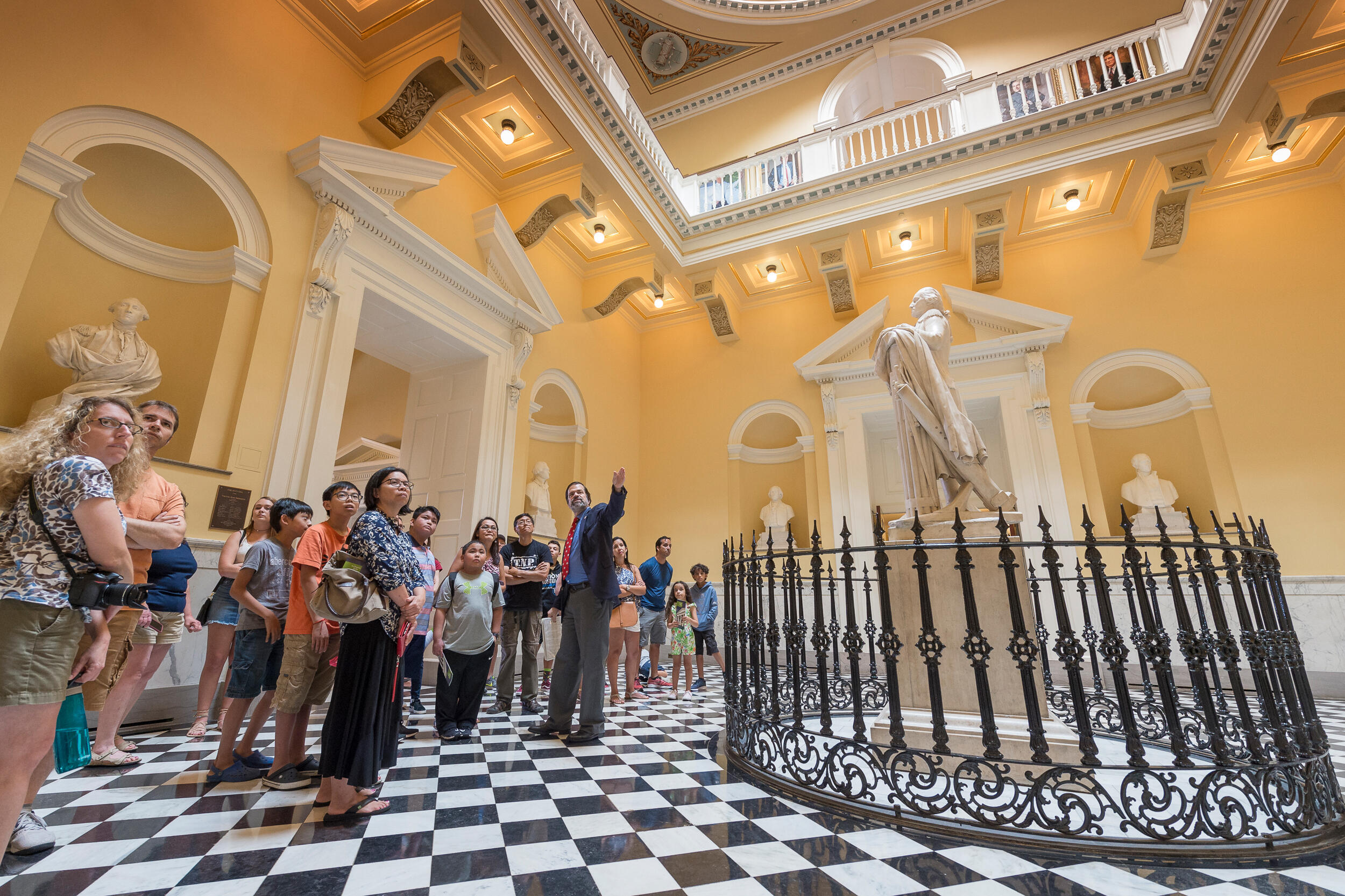 A tour guide takes a group of tourists past a statue of a historical figure and into an atrium at the Virginia State Capitol.