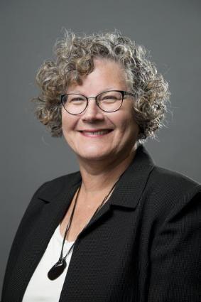 A portrait of a woman with short curly gray hair wearing a black sports coat, white shirt, and a necklace with a dark blue pendant. 