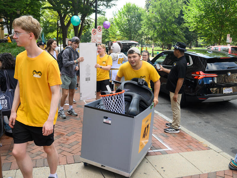 A photo of a sidewalk full of people. One man is pushing a bin filled with items he is moving into a dorm. 