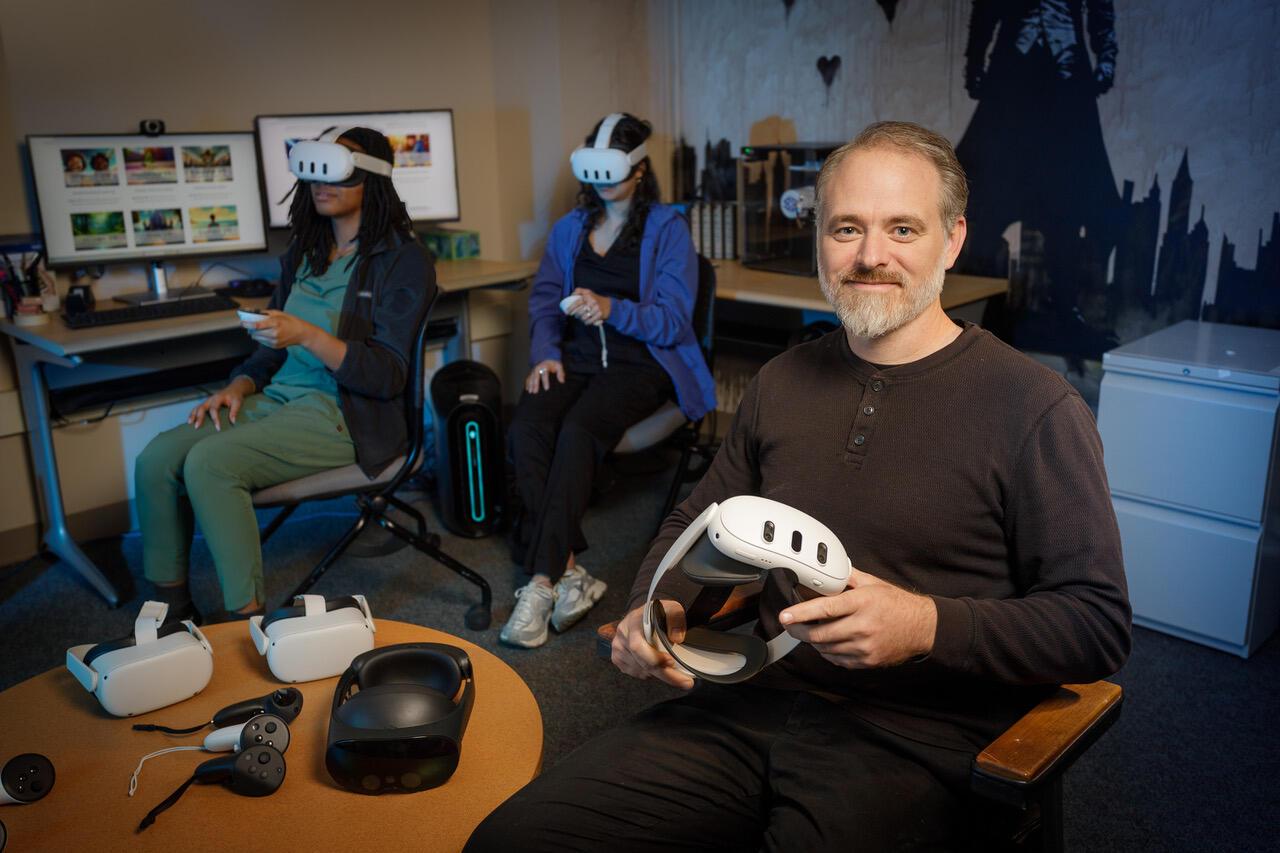 A photo of a man sitting in a chair holding a VR headset. Behind him are two women who are also sitting in chairs, but they are wearing VR headsets and holding VR controllers. On the table next to the man are three more VR headsets. 