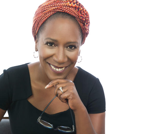 Woman in black shirt and red hair wrap smiling and holding eyeglasses.