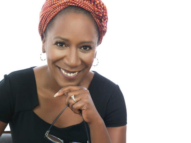 Woman in black shirt and red hair wrap smiling and holding eyeglasses.