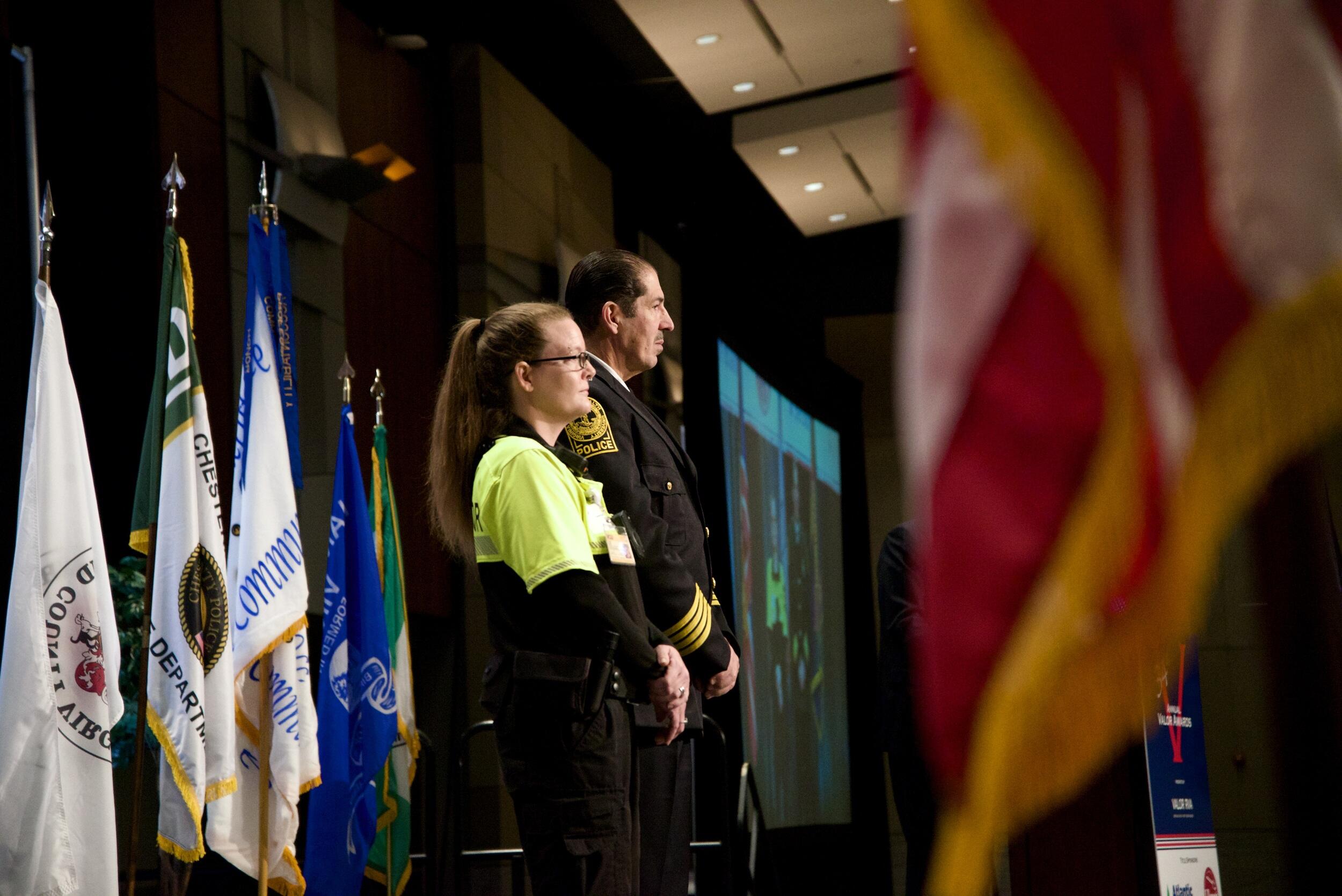 A photo of a man and woman standing on a stage with five flags on flag poles behind them. 