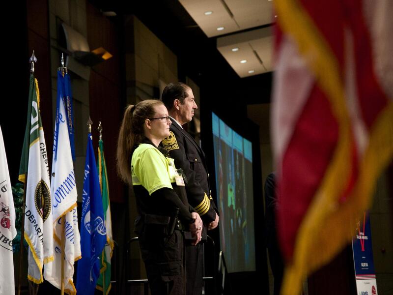 A photo of a man and woman standing on a stage with five flags on flag poles behind them. 