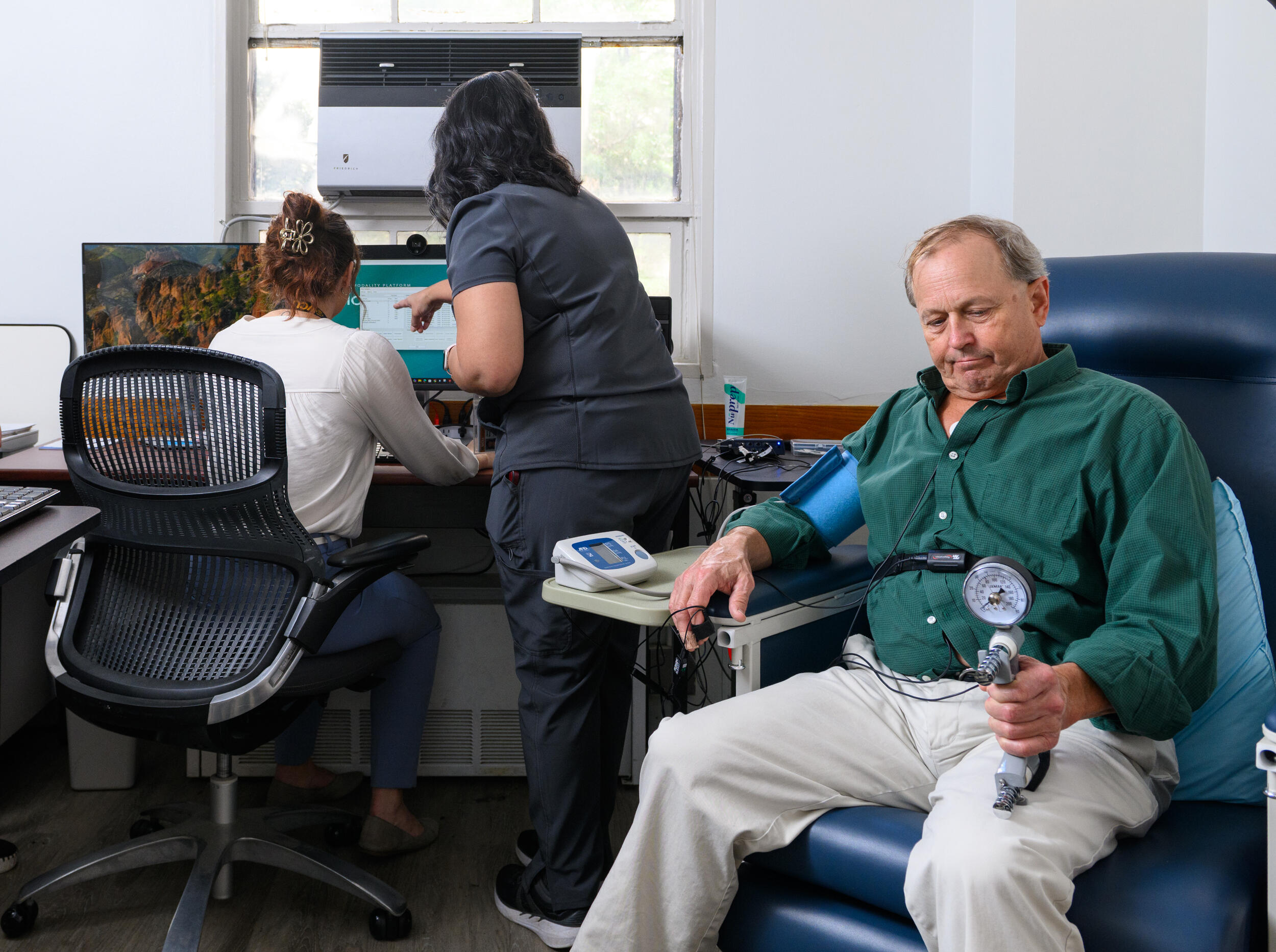 A photo of a man sitting in a chair and two women behind him at a desk. The woman on the left is sitting and the woman on the right is standing. 