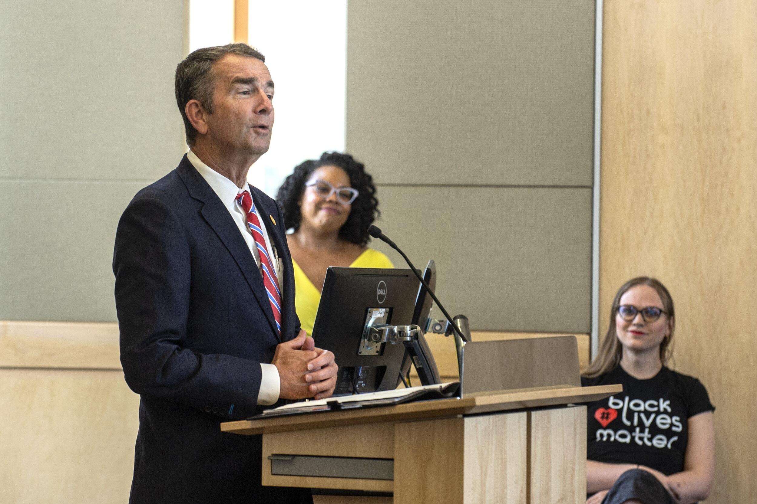 Gov. Ralph Northam speaking at a podium. Two conference attendees stand in the background.