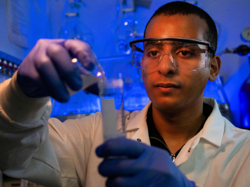 A man in safety goggles, gloves and a lab coat works in a lab.