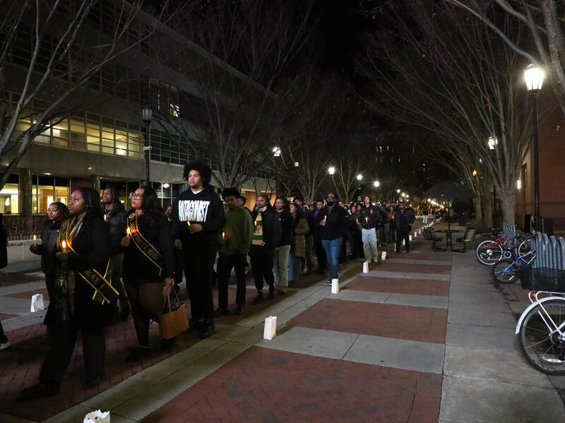 A photo of a line of people wearing all black walking in a line and holding candles. 