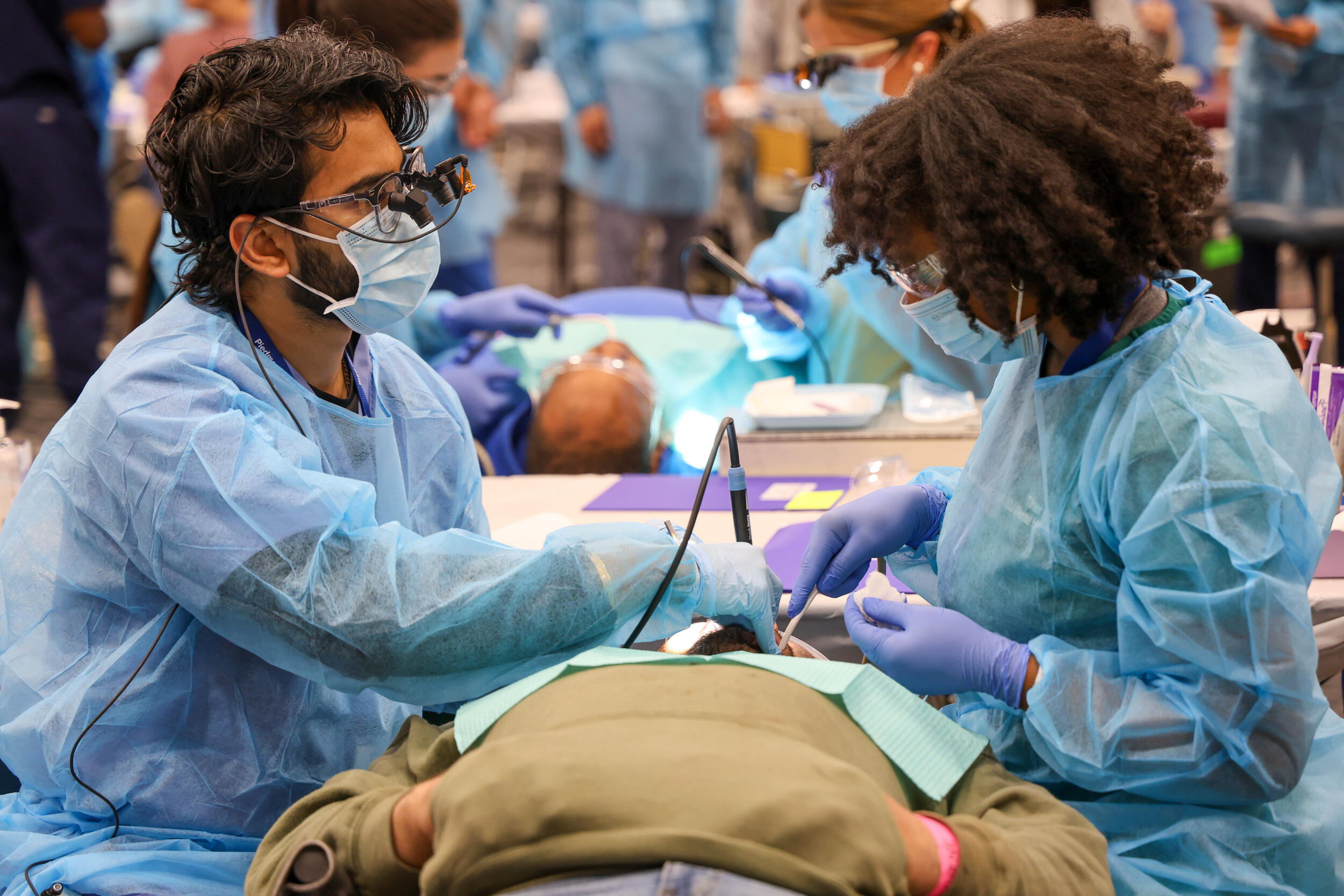 A photo of two people in blue dental scrubs cleaning the teeth of a patient lying down. 