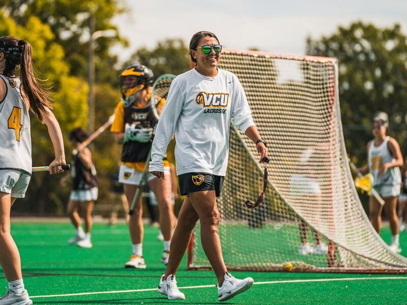A woman wearing sunglasses walking across a lacrosse field with other women holding sporting equipment behind her