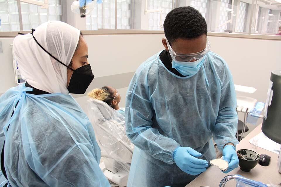 Two standing in a dental exam room in front of an exam chair with a woman in it. 