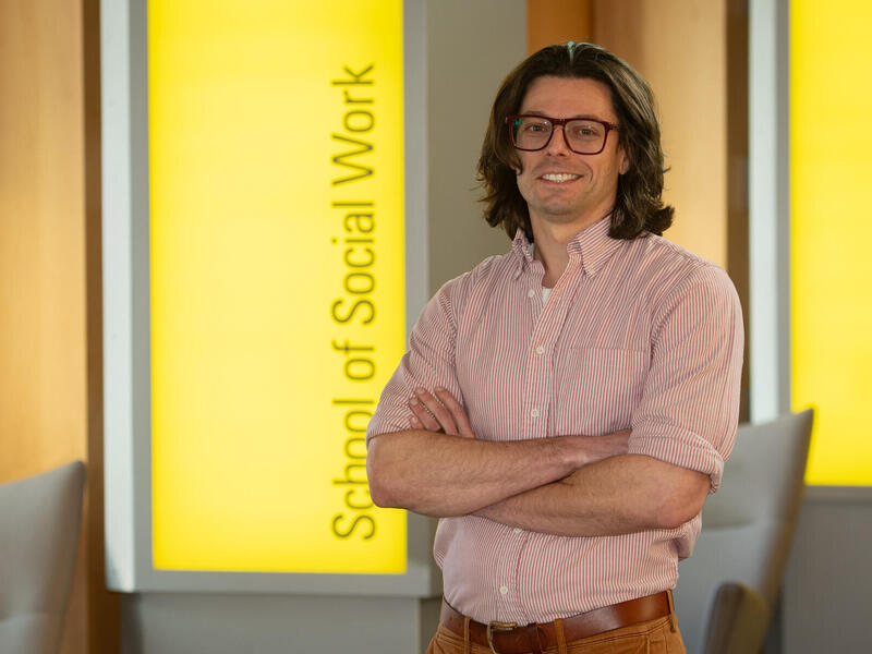 A photo of a man from the waist up. His arms are crossed against his chest. Behind the man is a yellow sign with black letters that says \"School of Social Work\"