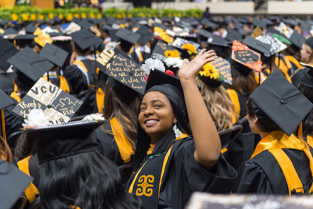 A graduating student seated among other graduating peers waves at a person.