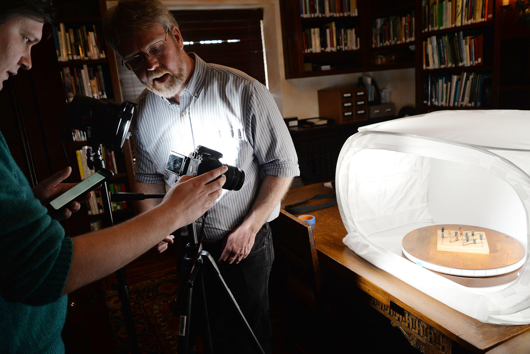 Two people aim a camera on a tripod at a wooden game in a light box.