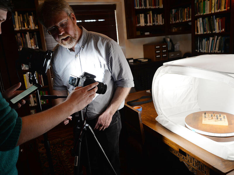 Two people aim a camera on a tripod at a wooden game in a light box.