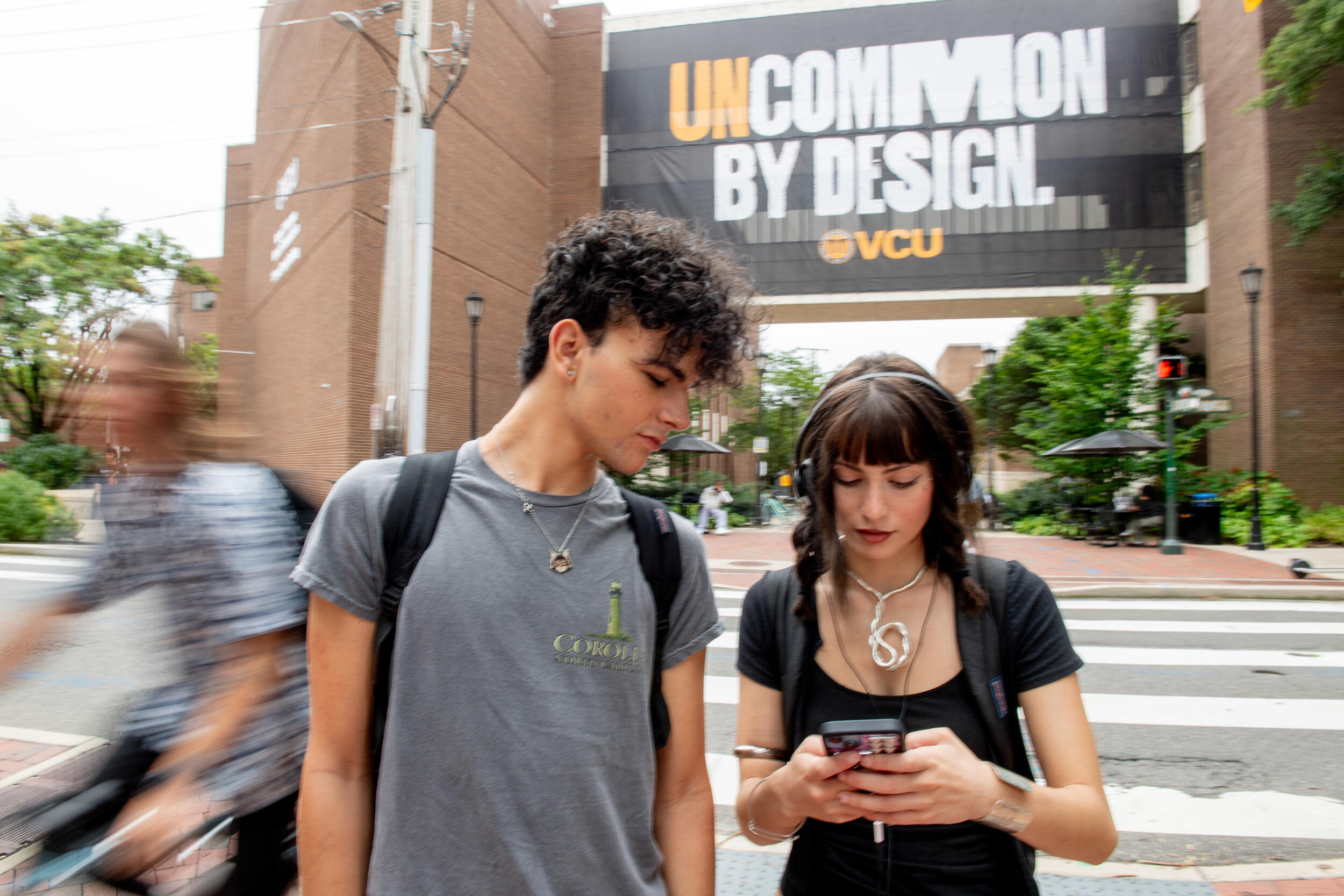 A photo of a man and a woman looking down at the woman's phone. Behind them is a banner that says \"UNCOMMON BY DESIGN VCU\" 