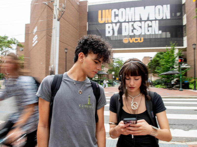 A photo of a man and a woman looking down at the woman's phone. Behind them is a banner that says \"UNCOMMON BY DESIGN VCU\" 