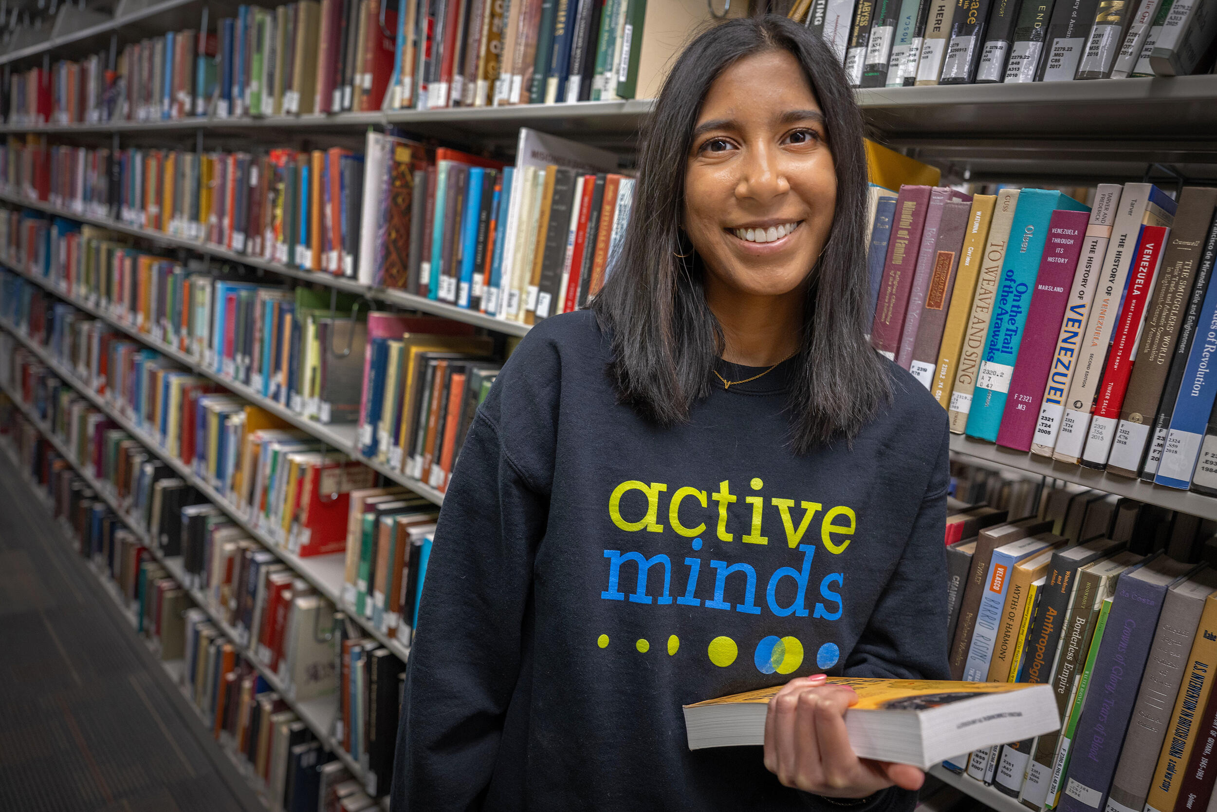 A photo of a woman from the waist up standing in front of library shelves covered in books. 