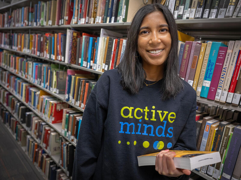 A photo of a woman from the waist up standing in front of library shelves covered in books. 
