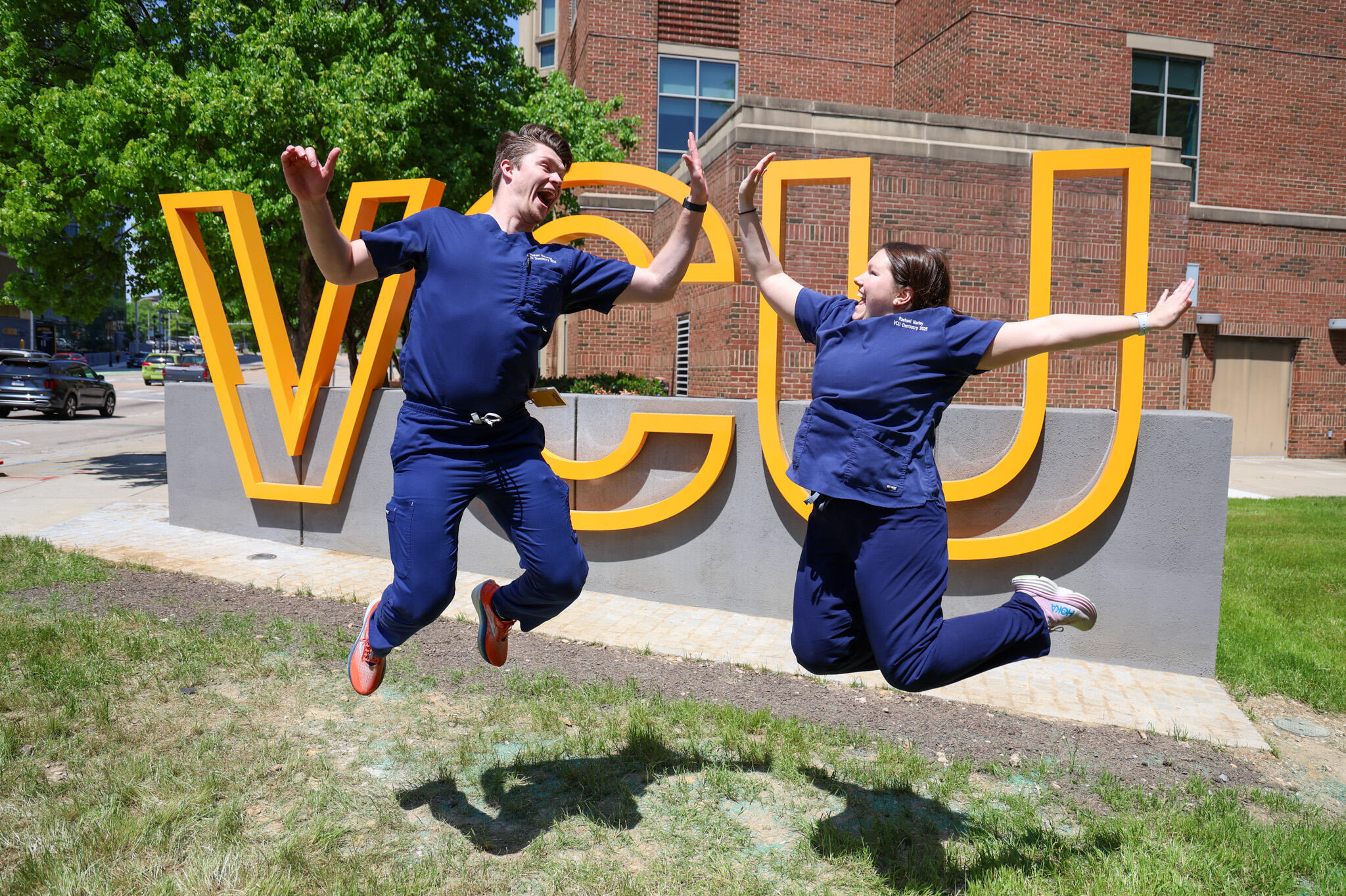 A photo of two people wearing blue dental scrubs jumping and high-fiving in front of a sign that says \"VCU\" in large yellow letters. 