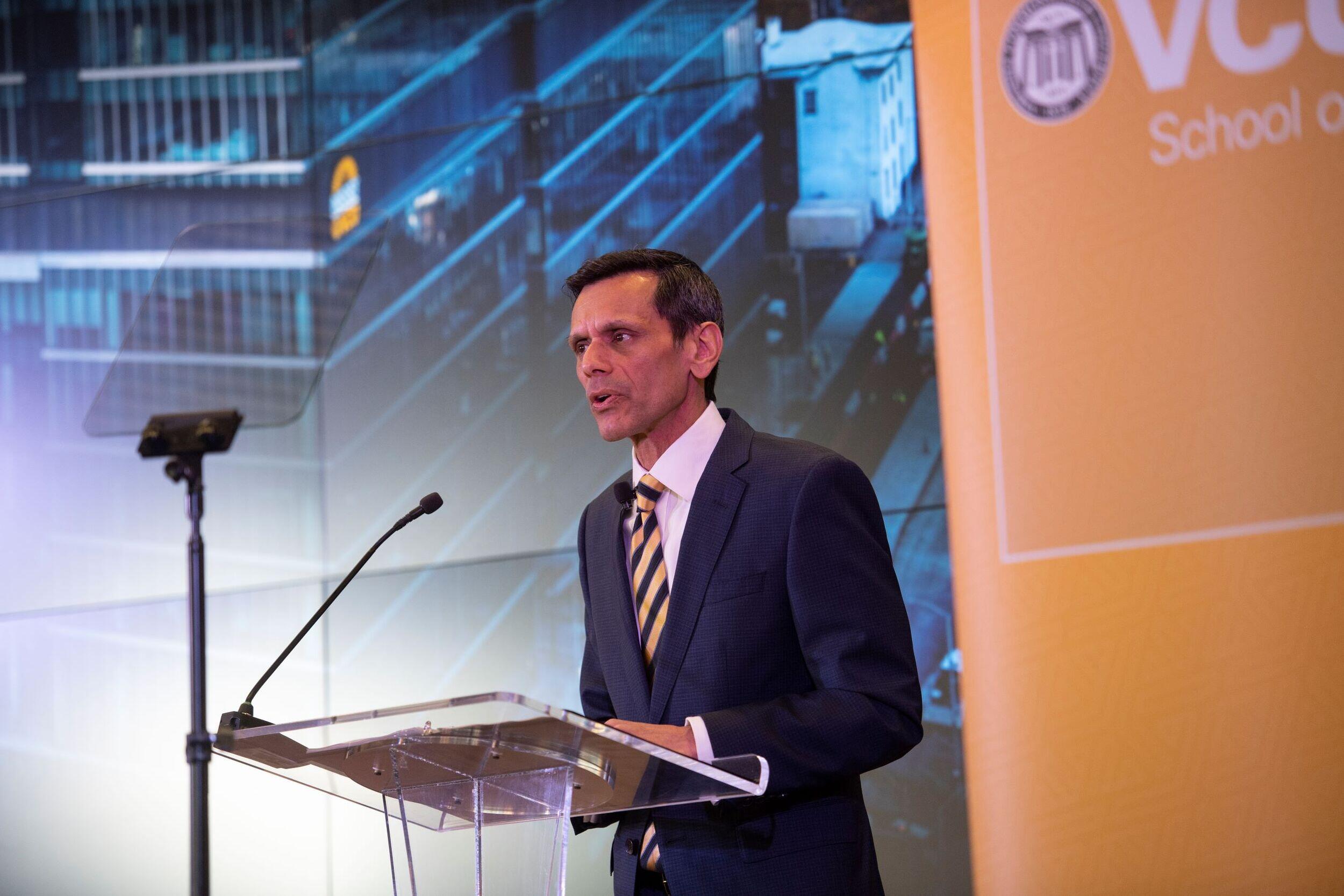 A man wearing a suit and tie speaking into a microphone at a podium made of clear plastic. 