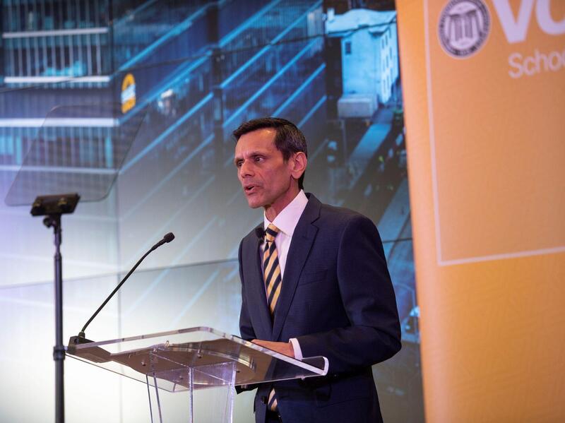 A man wearing a suit and tie speaking into a microphone at a podium made of clear plastic. 