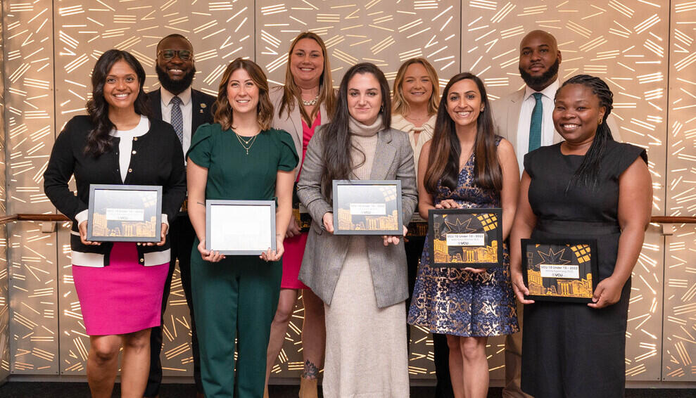 Nine people standing in a group. The five people standing in front are holding framed awards. 