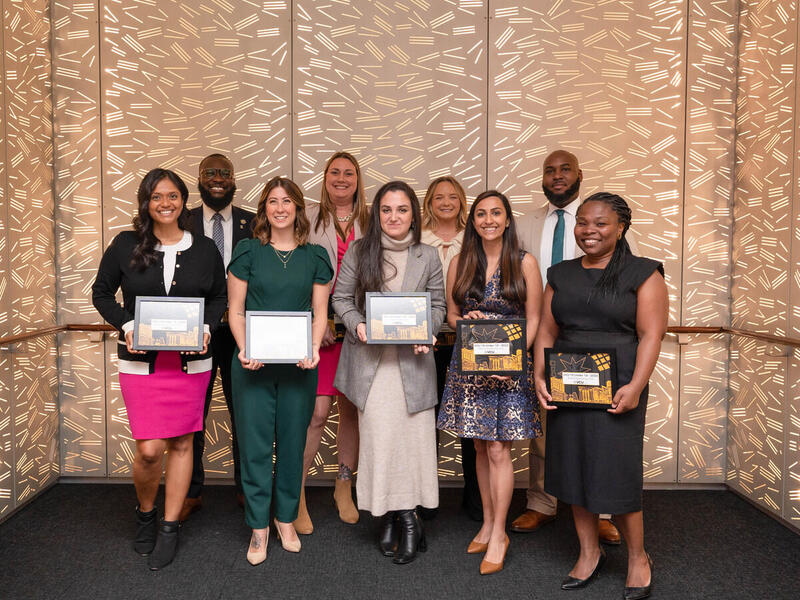 Nine people standing in a group. The five people standing in front are holding framed awards. 