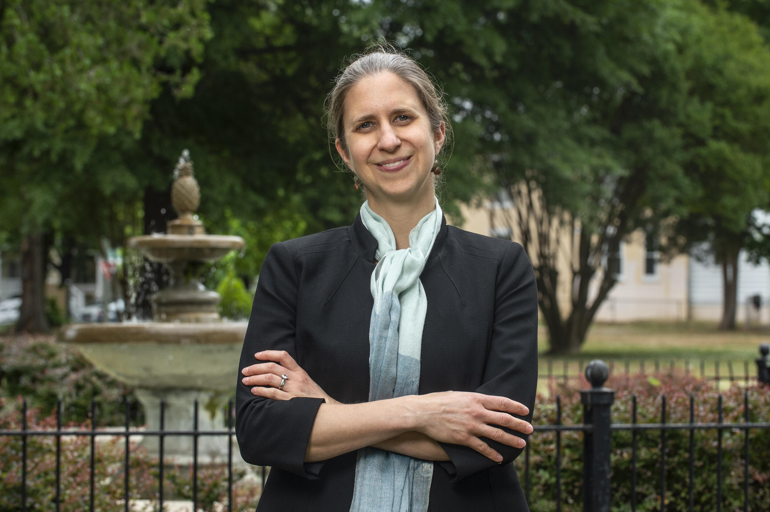 A photo of a woman from the chest up. She has her arms crossed against her chest. The woman is standing in front of a fountain in a park. 