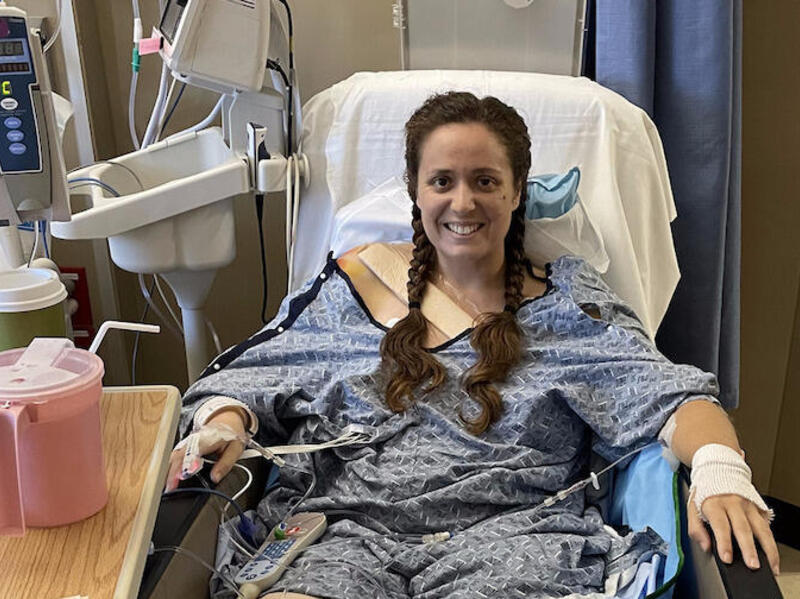 Natalie Rainer is sitting in a hospital bed smiling at the camera.