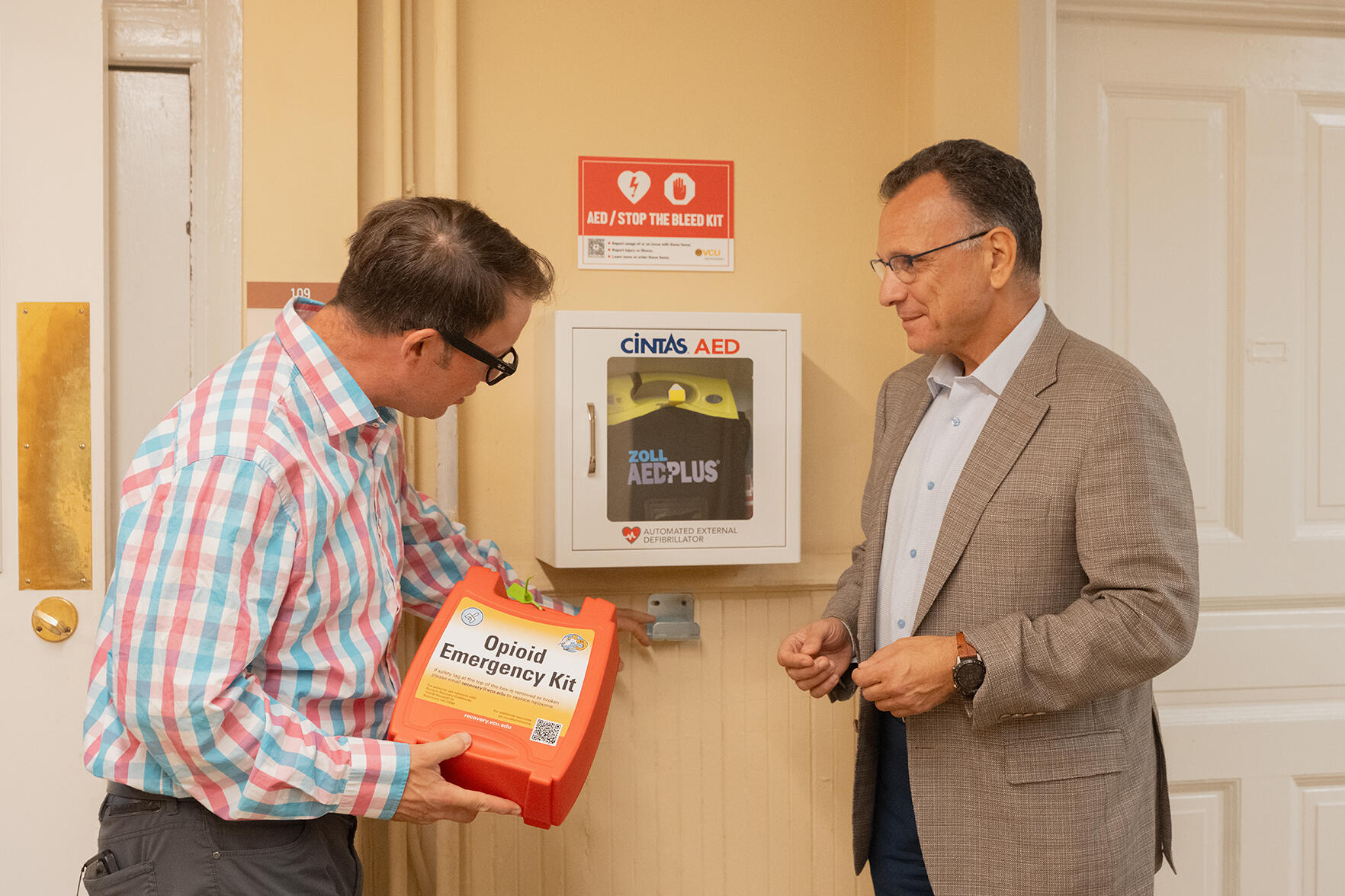 A photo of two men standing on either side of a defibrillator. The man on the left is holding an oragne box that says \"Opiod Emergency Kit.\"