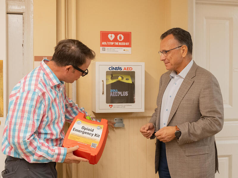 A photo of two men standing on either side of a defibrillator. The man on the left is holding an oragne box that says \"Opiod Emergency Kit.\"