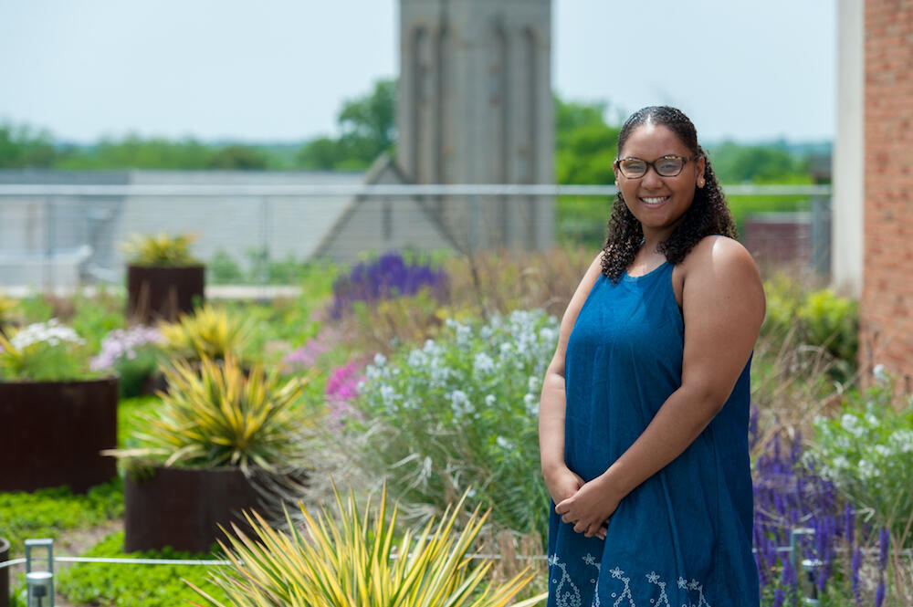 A smiling person in a blue dress stands in front of a rooftop garden.