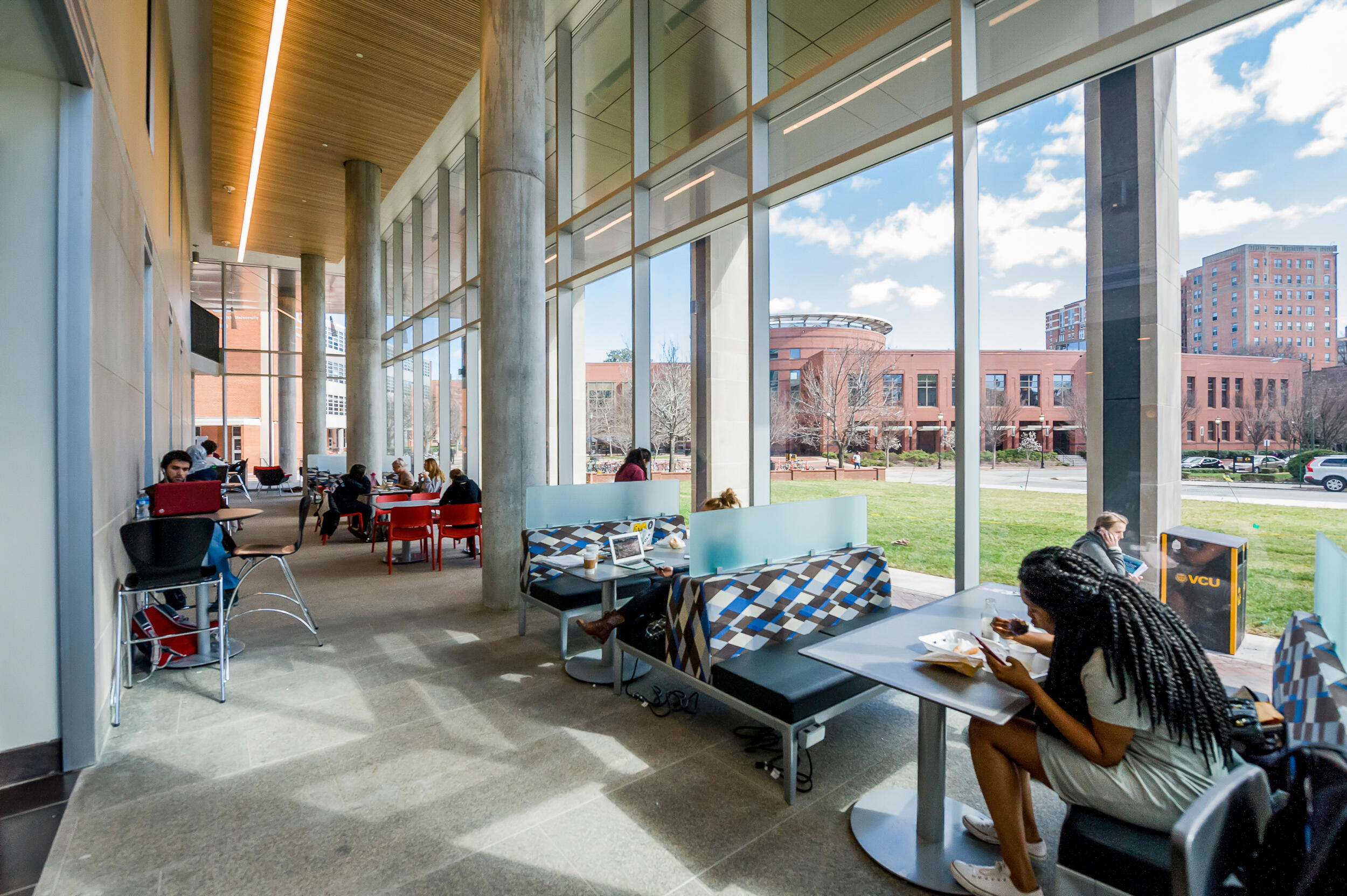 Students study by large windows on a sunny day at VCU Cabell Library across a green quad from Shafer Dining Court.
