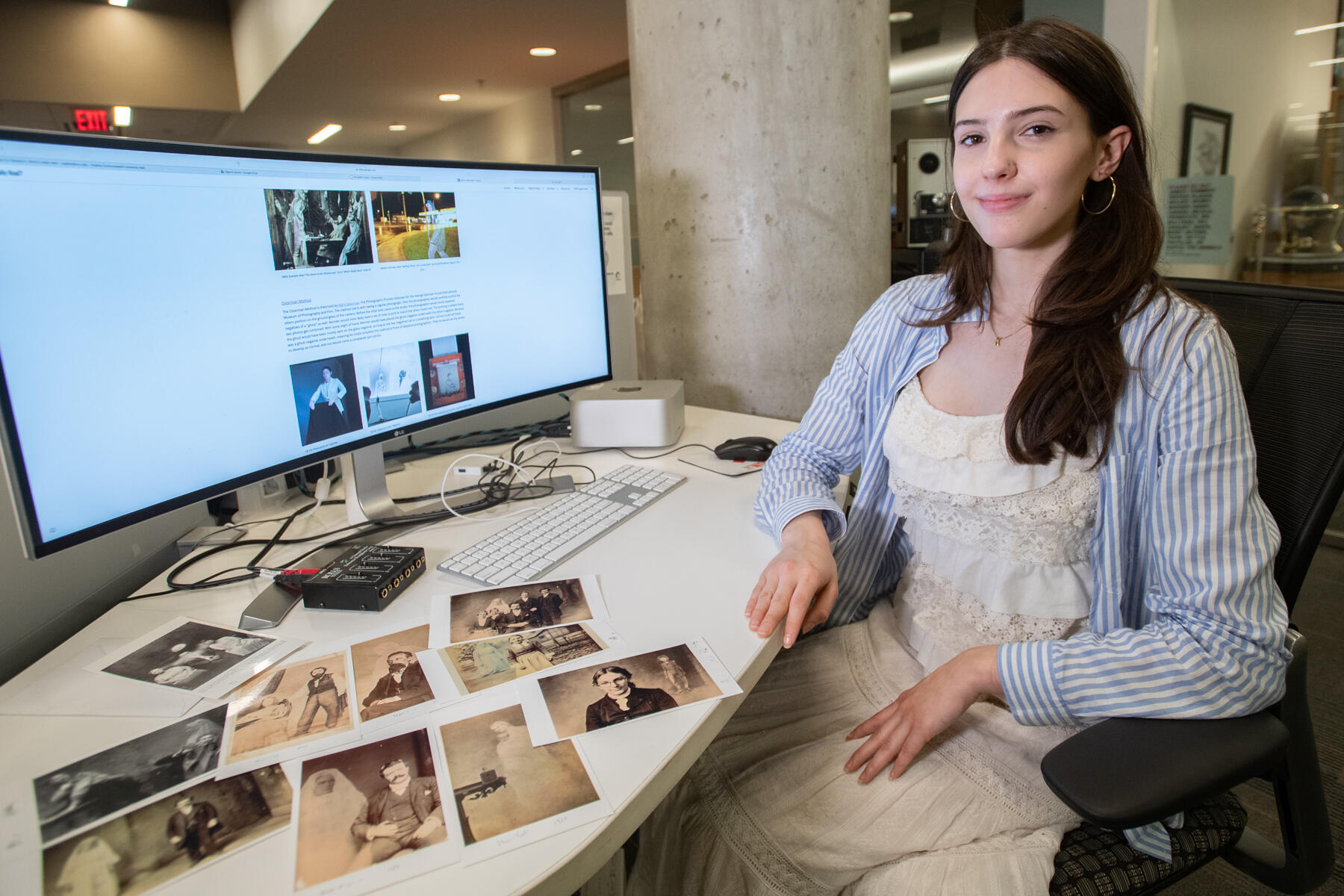A photo of a woman sitting in a chair next to a desk. The desk is covered in old photos and also has a computer monitor. On the computer monitor is illegible text and more old photos. 