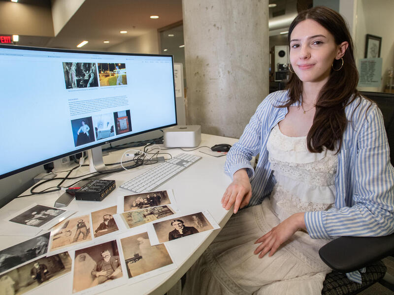 A photo of a woman sitting in a chair next to a desk. The desk is covered in old photos and also has a computer monitor. On the computer monitor is illegible text and more old photos. 
