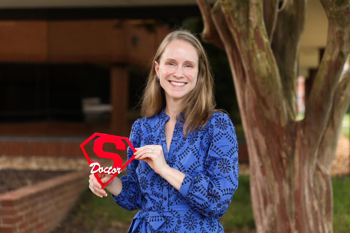 A photo of a woman from the waist up standing next to a tree. She is holding a sign with the super man logo with the word \"Doctor\" written overtop in white letters. 