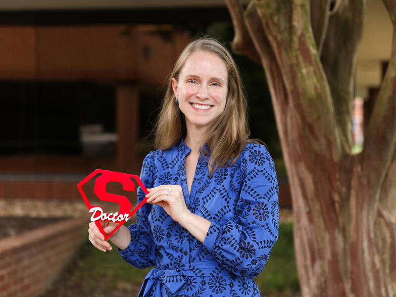 A photo of a woman from the waist up standing next to a tree. She is holding a sign with the super man logo with the word \"Doctor\" written overtop in white letters. 