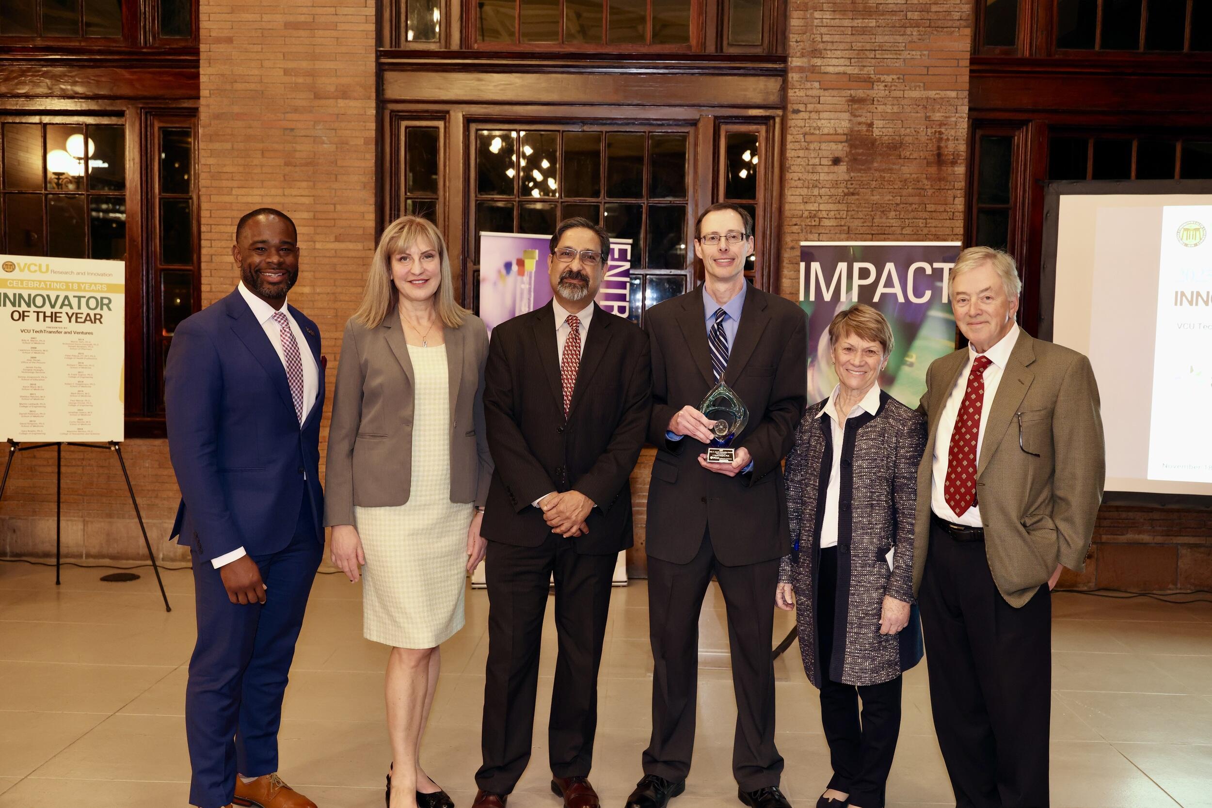 A group photo of six people standing. The third man to the right is holding a glass trophy. 
